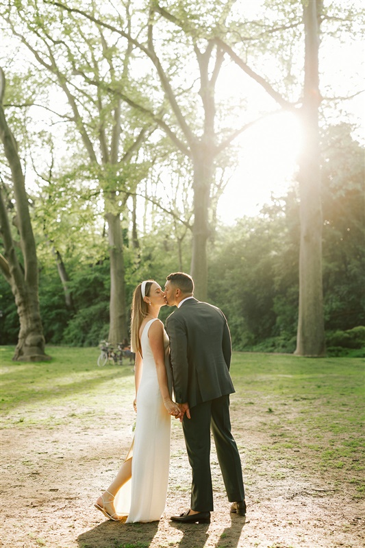 Bride and groom sharing a kiss while walking through Prospect Park with their backs turned toward the camera