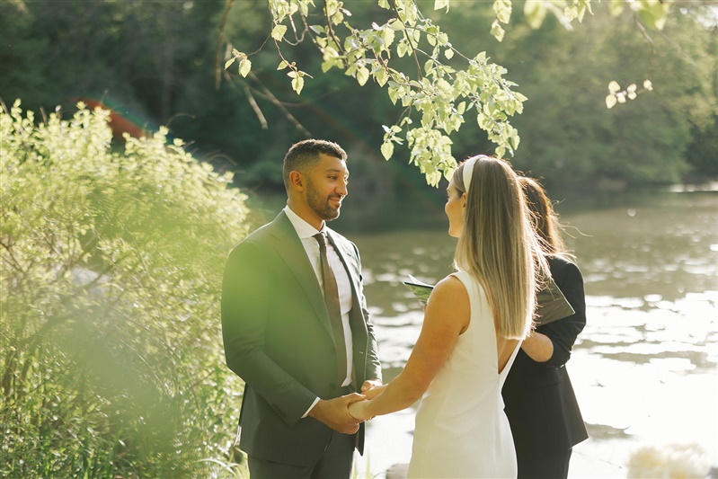 Photo of groom smiling at brides while they are holding hands during their waterfront ceremony