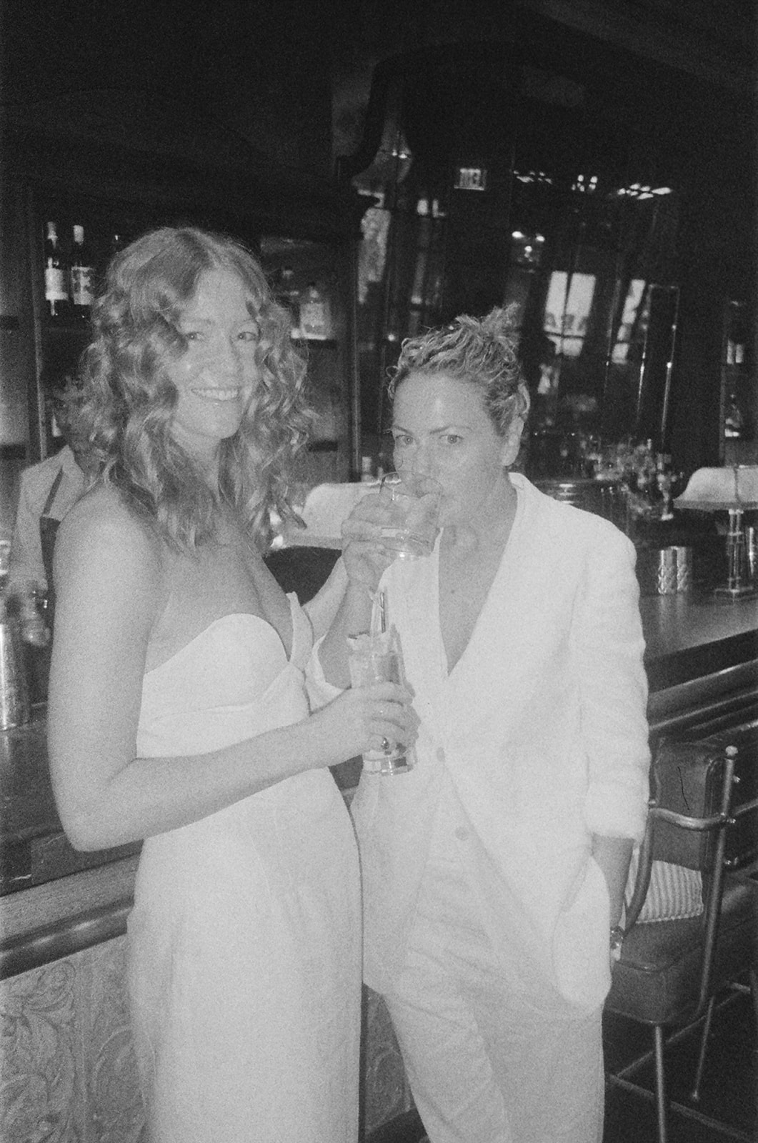 Wedding photos at a bar with one bride drinking a drink and the other standing holding a drink
