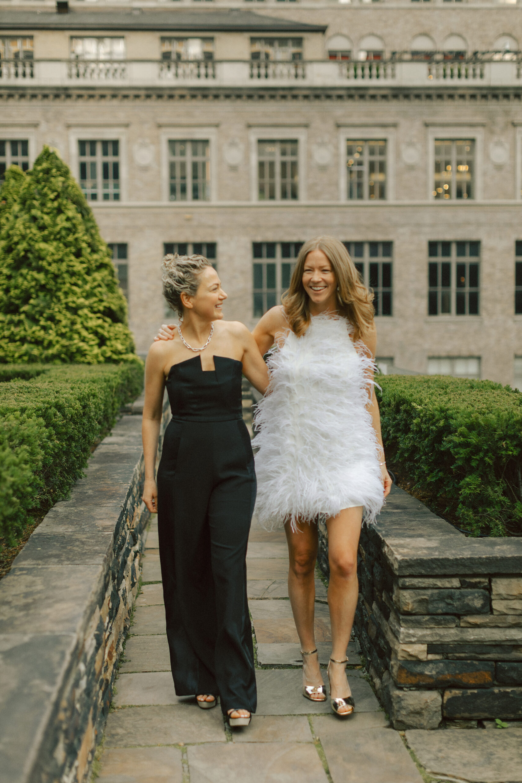 Couple walking toward the camera, one in a black jumpsuit and the other in a feather mini dress