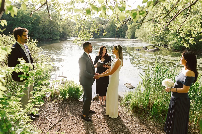 Waterfront wedding ceremony in Prospect Park, surrounded by trees and sunlight with bride and groom holding hands while officiant speaks.